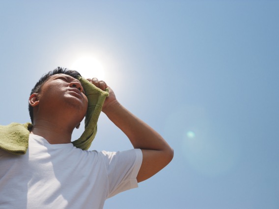 Looking up at a young man in a white t-shirt as he dabs sweat from his brow with a green towel, the sun glaring behind his head.