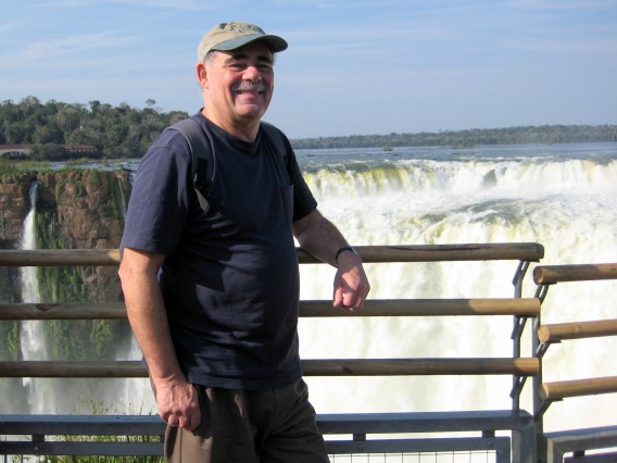 Bob leans with one arm against a metal railing in front of a waterfall wearing a brown baseball cap, navy blue shirt and brown pants.