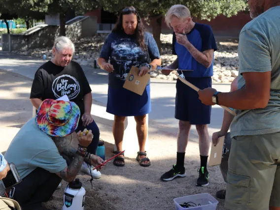 A man in a tie-dye bucket hat kneels over a whole dug in the dirt as a circle of teachers with clipboards look on.