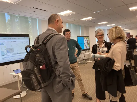 Egoitz Laparra stands with his hands on his hips in a green sweater and slacks as Laura López-Hoffman, in a black suit, speaks with two EPA representatives at the White House Summit. The walls of the room feature several screens demoing software.