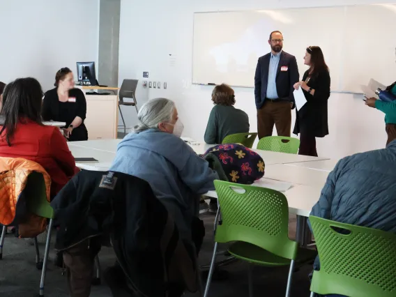 Ladd Keith and Leslie Ethen stand at the front of a classroom with several workshop participants seated in front of them at white tables in green folding chairs. 