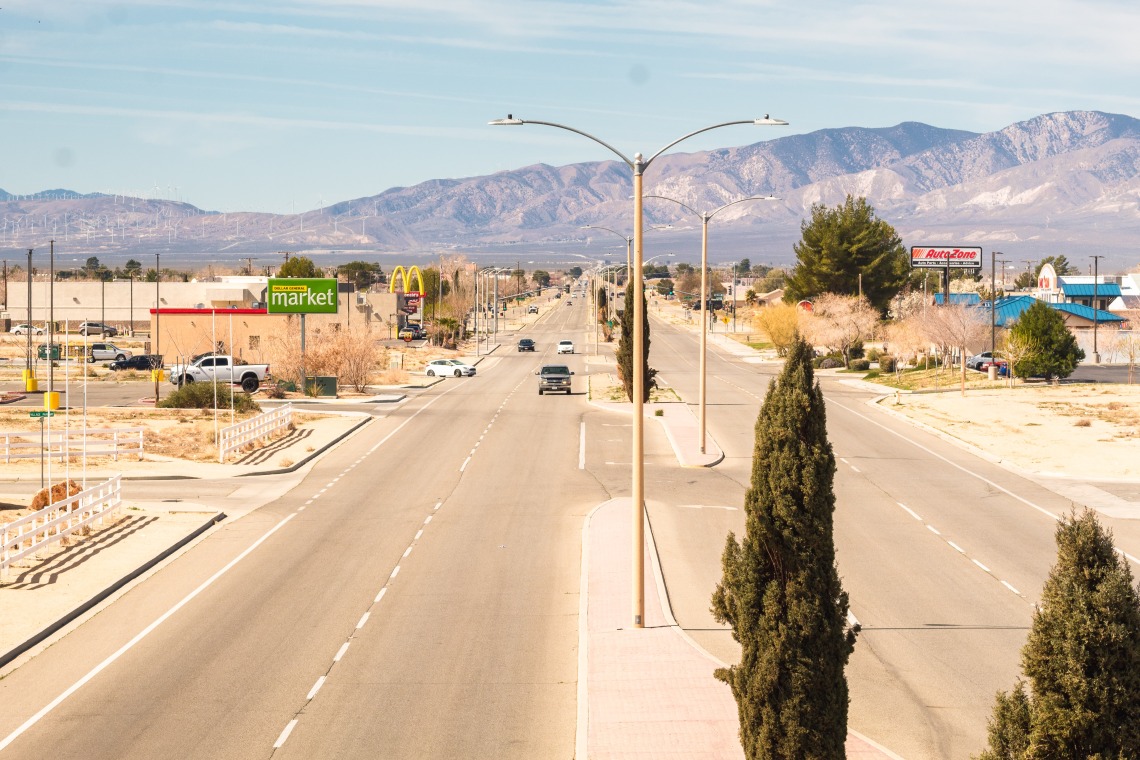 A photo of a paved desert street with a few cars, sparse trees, and mountains in the background against a pale blue sky.