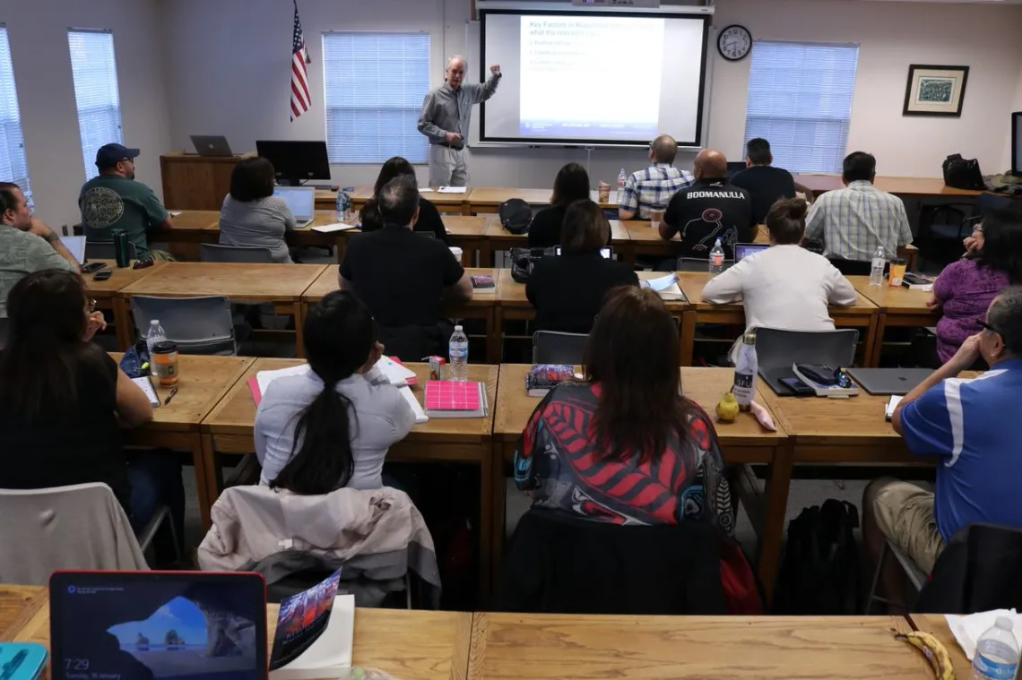 A view of a full classroom from the back row with Dr. Stephen Cornell gesturing near a projector screen at the front of the room