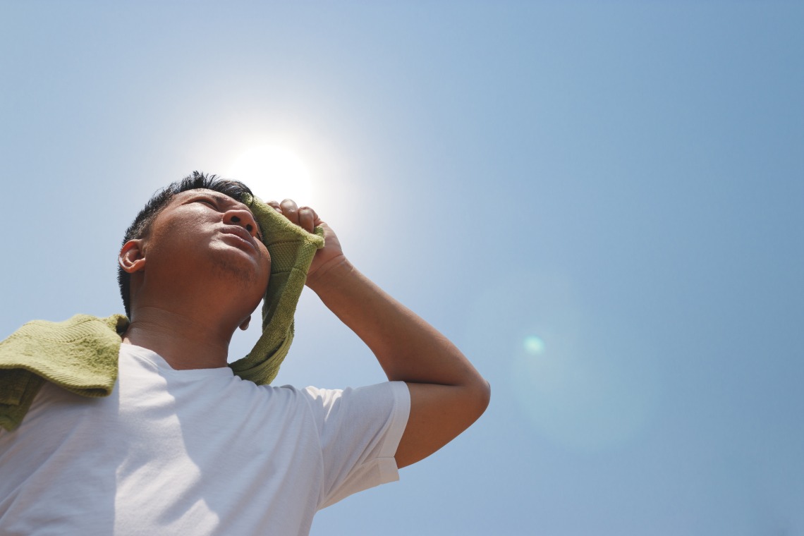 Looking up at a young man in a white t-shirt as he dabs sweat from his brow with a green towel, the sun glaring behind his head.