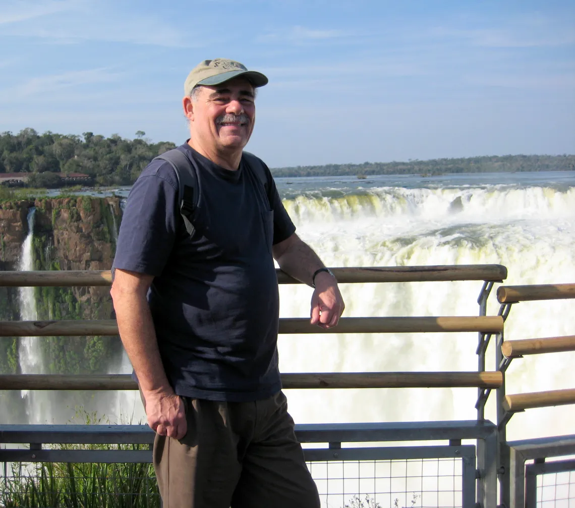 Bob leans with one arm against a metal railing in front of a waterfall wearing a brown baseball cap, navy blue shirt and brown pants.