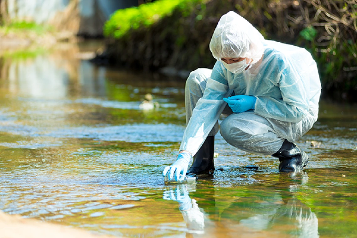A person in full body PPE kneels in a stream to collect water in a test tube
