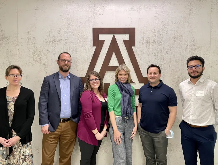 Six people dressed in business attire smile in front of the University of Arizona logo