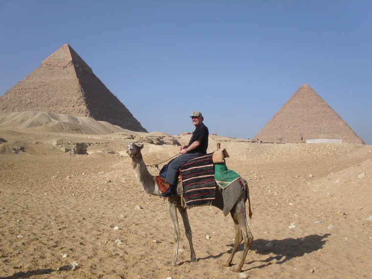 Bob in a black t-shirt, brown cap and gray pants sits atop a camel adorned with a striped blanket and saddle in from of the Pyramids of Giza.
