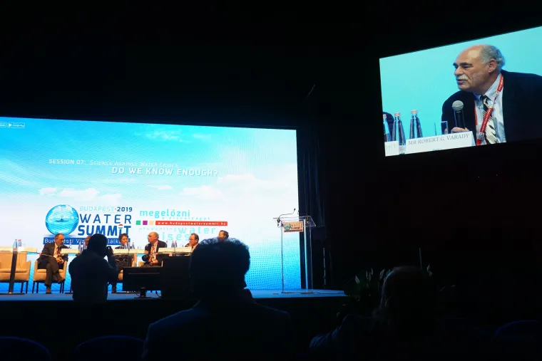 A monitor to the upper right of the image shows Bob Varady in a suit speaking to a microphone. The stage with panel members including Bob appears at the bottom left of the image.