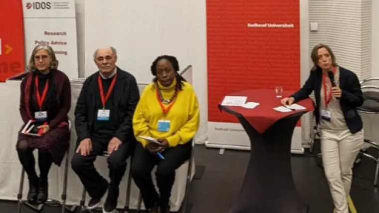 Panelists sit in tall chairs on the left as the moderator leans on a high-top table on the right side of the screen. A red and white IDOS banner  stands upright behind the speakers in the top left corner of the image.