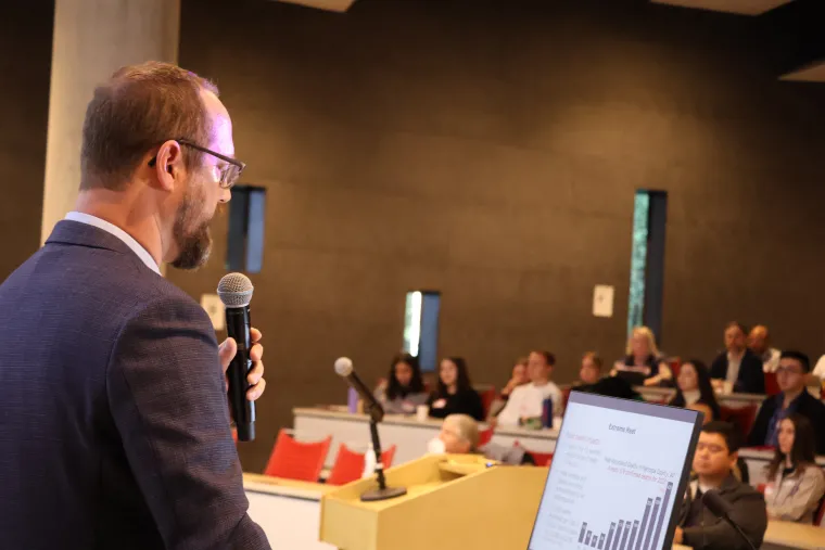 Ladd Keith speaks into a microphone as a crowd listens in from the auditorium. Photo shows Keith in profile as he looks at a slide show displaying a graph on the monitor in front of him.