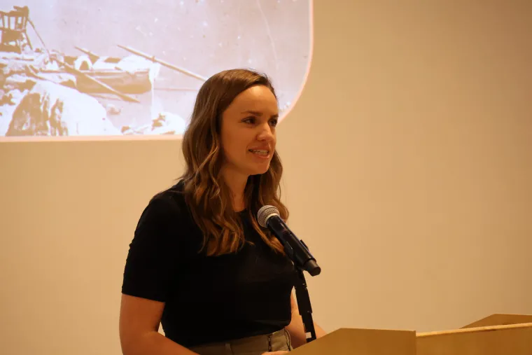 Gina Gilson speaks from a podium in a black polo shirt.