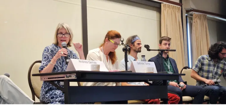 Andrea Gerlak speaks into a microphone while seated next to four other panelists to her left.