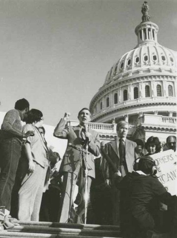 A man in a suit speaking into a microphone in front of a state capitol building