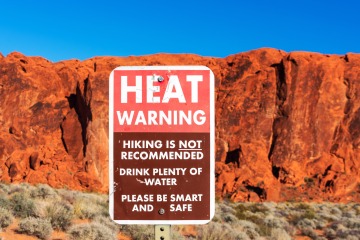 An image of a sign stating "Heat Warning: Hiking is not recommended. Drink plenty of water. Please be smart and safe." with red desert mountain cliffs in the background.