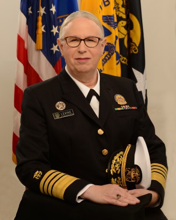 An admiral in uniform seated in front of the U.S. and military flags.