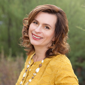 A portrait of Professor Jamie Edgin smiling outdoors in a yellow blouse and large beaded necklace.