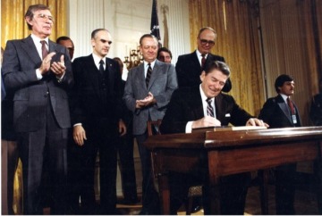 A color image of President Reagan signing a bill into law with seven lawmakers behind him, including Mo Udall who stands clapping at the far left of the image.