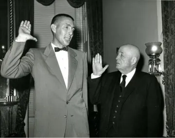 Udall in a bow tie is sworn in by Speaker of the House Sam Rayburn, who is significantly shorter than Udall. Both men have their right hands raised in the image.