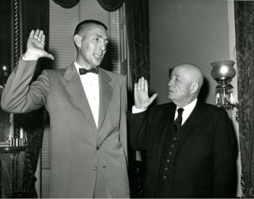 Udall in a bow tie is sworn in by Speaker of the House Sam Rayburn, who is significantly shorter than Udall. Both men have their right hands raised in the image.