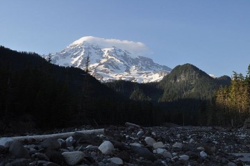 A snow-capped mountain rises above hills covered with evergreen trees with a rocky creek bed in the foreground.