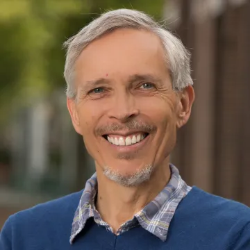 Outdoor portrait of Impey wearing a blue sweater over a blue plaid collared shirt in front of a blurry backdrop.