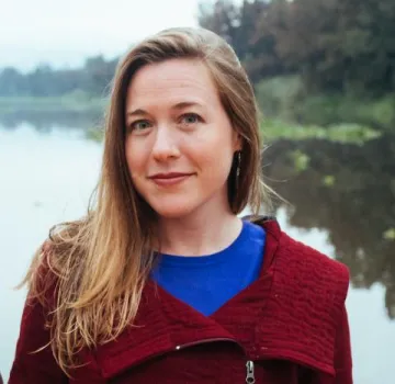 Portrait of Tellman in a red sweater with an oversized collar over a blue shirt with a lake in the background.