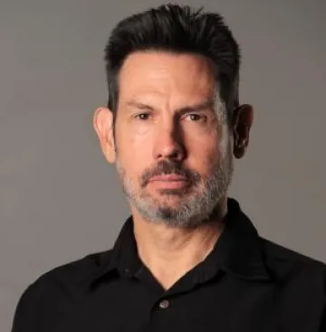 Portrait of a stoic Mulcahy in a black polo shirt against a gray background.