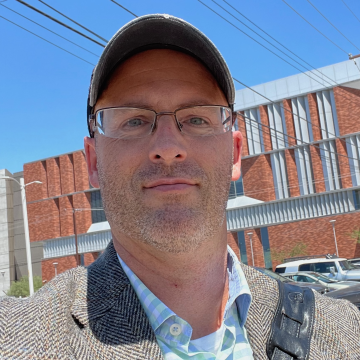 A portrait of Austin Duncan in front of powerlines and a red brick building. He wears wireframe glasses, a ball cap, a tweed jacket and blue and white striped shirt. The strap of a bag is visible on his left shoulder.