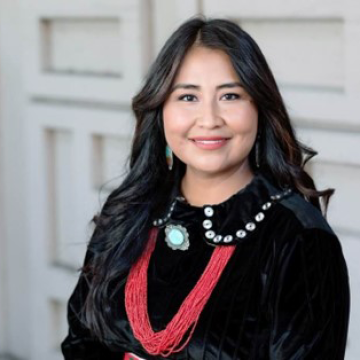 A portrait of Cherie De Vore in front of a white door in a black velvet blouse with white embellishments on the collar. She wears a large silver and turquoise pendant with a multi-strand coral bead necklace.