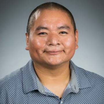 A portrait of Darryl Reano in a blue collared shirt with a white polka dot pattern against a gray background.