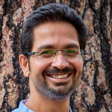 A portrait of Pranay Ranjan in front of the broad trunk of a pine tree. He wears frameless square glasses and a short beard.