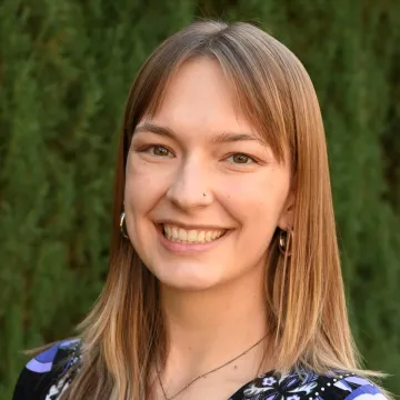 Ms. Broski smiles in a blue floral print blouse in front of foliage.