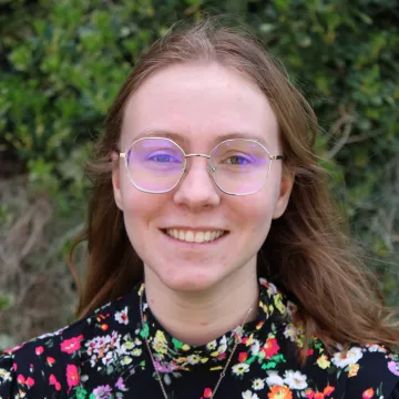 Ms. Clark smiles in wire-framed glasses and a black floral print blouse in front of foliage.