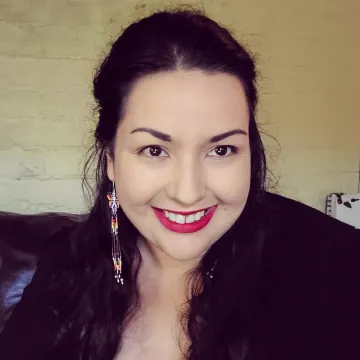 Ms. Kouyoumdjian smiles in a black coat and colorful dangling earrings indoors in front of a white brick wall.