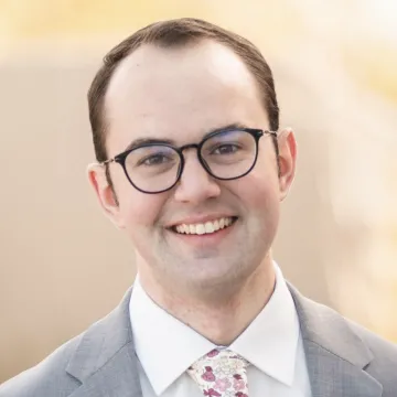 Mr. Macy smiles in black frame glasses, a gray suit, white shirt and colorful tie in an outdoor setting.