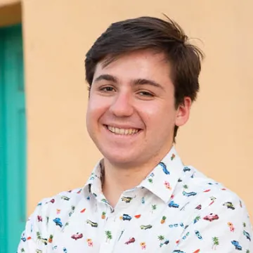 Mr. Pongratz smiles in front of a beige wall and green door in a white button-up shirt with colorful beach iconography on it.