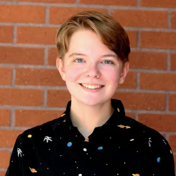 Mr. Rose smiles in front of a red brick wall wearing a black button-up shirt emblazoned with space iconography