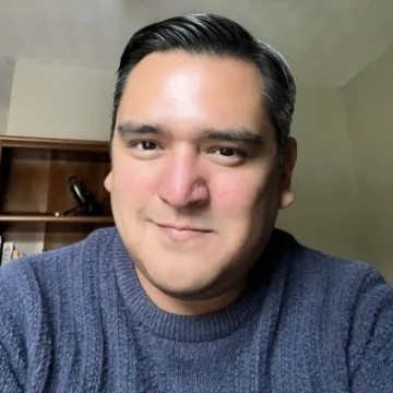 Tory Fodder smiles into the camera in a blue sweater. A dark wooden bookshelf can be seen over his right shoulder (left side of the image).