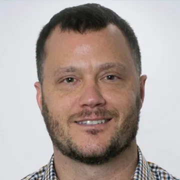 Jason Jurjevich smiles in a multi-colored plaid shirt against a gray background.