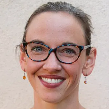 Robin Reinke smiles in multi-color-framed glasses against a white stucco wall.