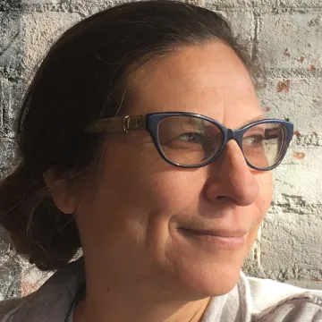 Elaine Romero wears a white sweater and smiles in black-framed cat-eye while peering off to one side in front of a concrete block wall.