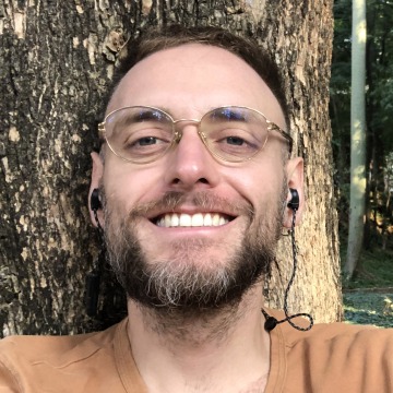 A selfie of Daniel Mendizabel Castillo in an orange t-shirt and ear buds taken in front of a tree trunk.