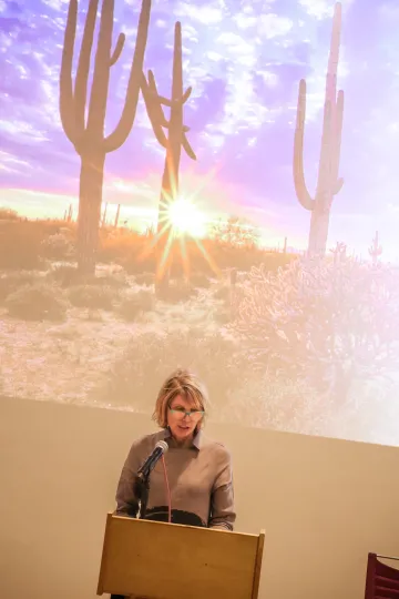 Andrea gerlak speaks behind a podium in front of a sunset behind saguaro cacti projected on the wall behind her.