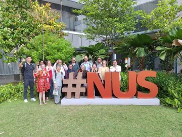 GHHIN members pose by large red letters reading "#NUS" on the Yale-National University of Singapore campus outside a gray building on a grass lawn.