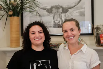 Caitlin Brady and Gina Gilson pose in front of a photo of a steer above a fireplace in the Udall Center lobby.