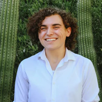 Jack Miller poses in a white collared shirt in front of some greenery and two columnar cacti.