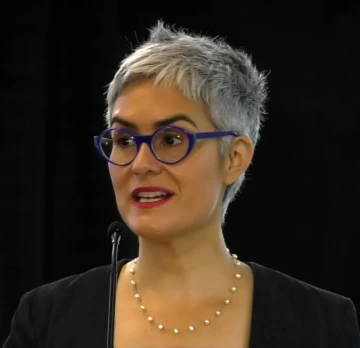 Laura López-Hoffman speaks into a small black microphone in front of a black background in a black top, gold necklace and blue-framed glasses.