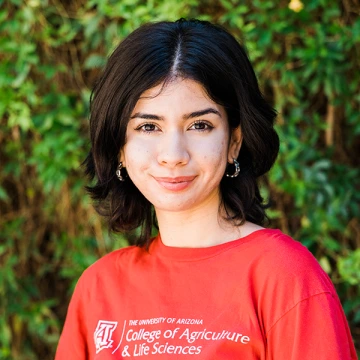 Illiana Samorano flashes a close-lipped smile in a red College of Agriculture t-shirt while standing in front of foliage