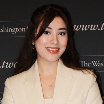 Malika Tuychiboeva smiles in front of a black step-and-repeat banner with white text while wearing a light tan suit